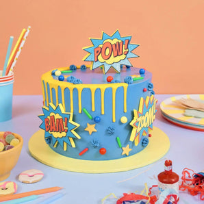 Comic Book Superhero Cake with blue frosting, yellow drip, and comic-style POW!, BAM!, and ZOOM! decorations by Crumbs & Doilies